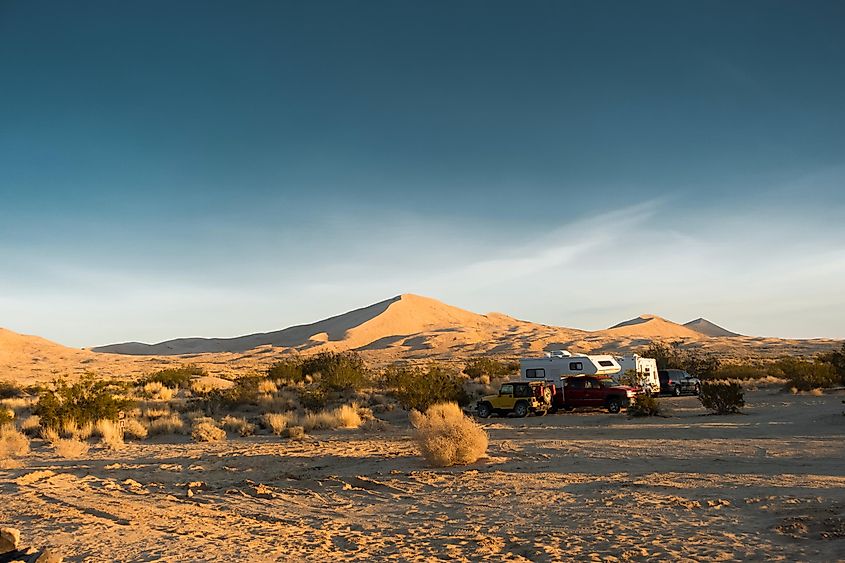 The main dune of the Kelso Dunes is seen at sunrise. Campers and recreational vehicles, RVs, cars, trucks, SUVs, are seen in the foreground. Mohave National Park, California