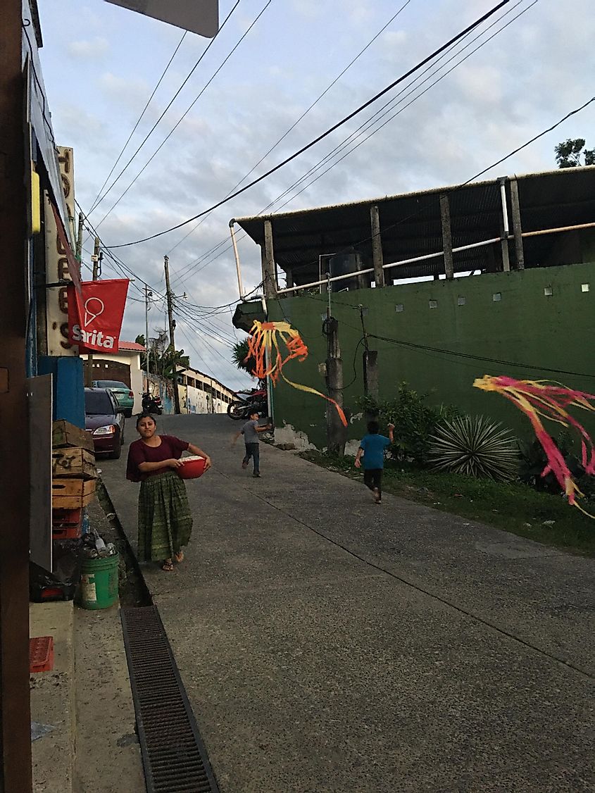 Two kids flying kites in the street while a woman carries items to the store.