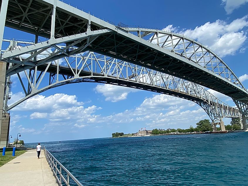 A man walks a riverside path underneath a pair of massive steel bridges.