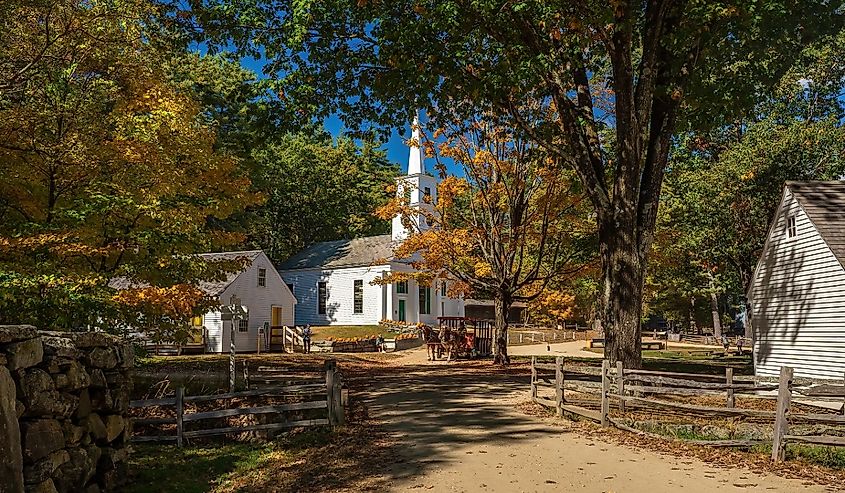 Historic village scene with a white church and horse-drawn carriage amid vibrant autumn trees. A stone fence lines the sandy path under a clear blue sky.