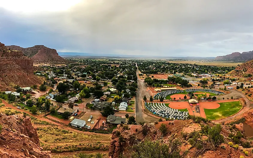 Aerial view of a small town nestled in a desert landscape with red rock formations. The town features scattered homes, trees, and a circular park. The sky is overcast.