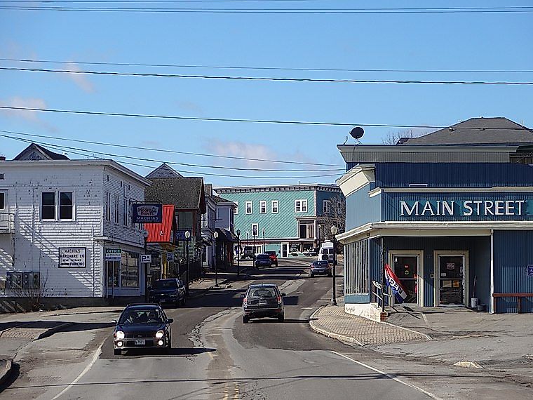 A small-town street scene with cars on the road and blue sky above. The blue building on the right has a "MAIN STREET" sign, conveying a quiet, quaint vibe.
