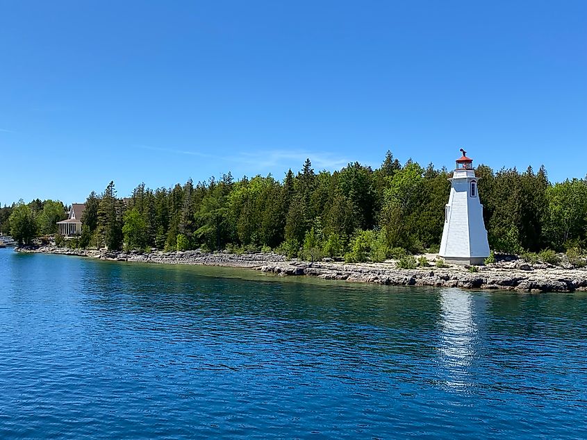 A white lighthouse with red trim stands at the mouth of a small, tree-lined harbour