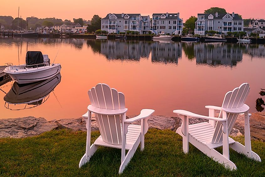 Two white Adirondack chairs on the beach, a moored boat, and smoke from a wildfire over Mystic River marina village, Connecticut.