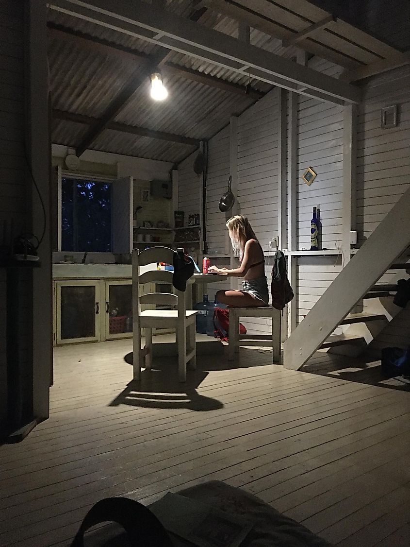 A young woman works at a table under a single light bulb inside of a jungle cabin at night.