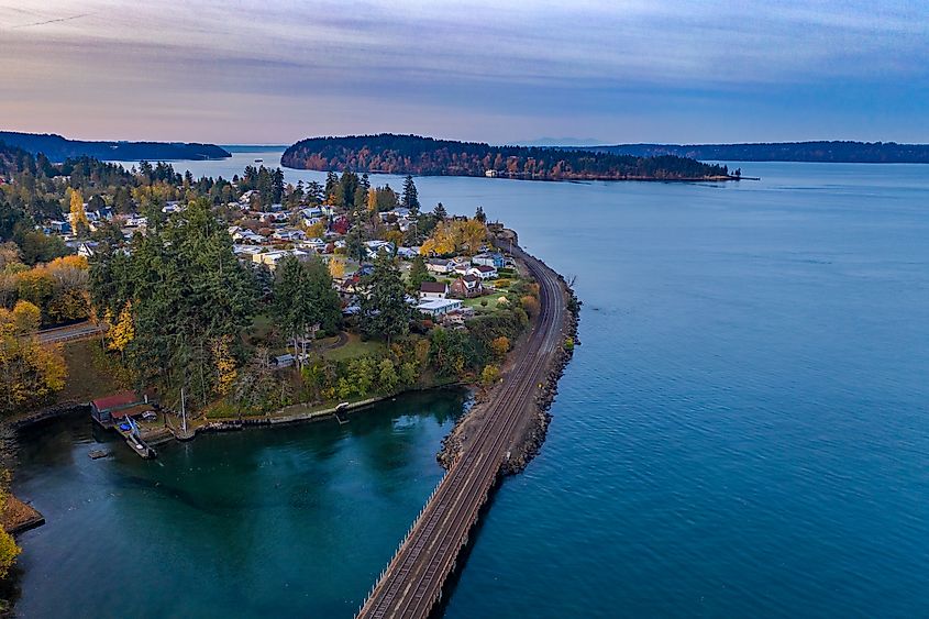Aerial view of a serene coastal town with autumn trees and quaint houses near the water's edge. A rail line curves through the scene, bordered by calm blue waters and distant islands under a soft pastel sky.