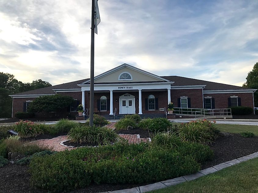 Brick town hall with white columns and arched windows, surrounded by landscaped gardens under a blue sky. A flagpole stands in front.