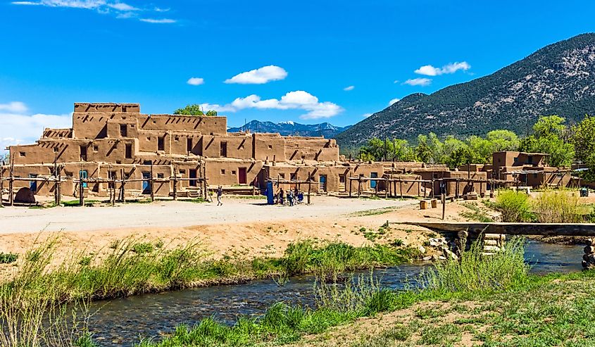 Pueblo dwellings in New Mexico with mountains in the background and a river in the forefront.