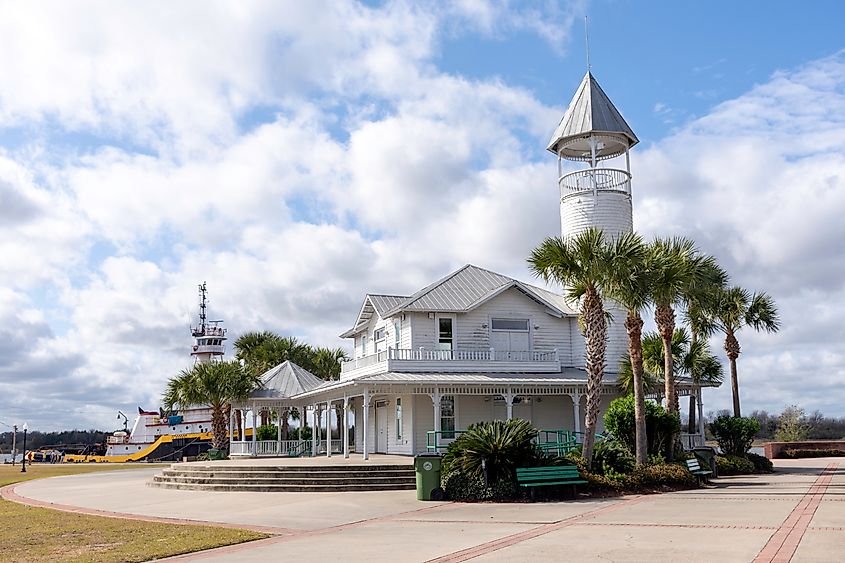 A white, two-story building with a conical-roofed tower sits beside palm trees under a cloudy sky. A colorful ship is in the background, evoking a coastal feel.