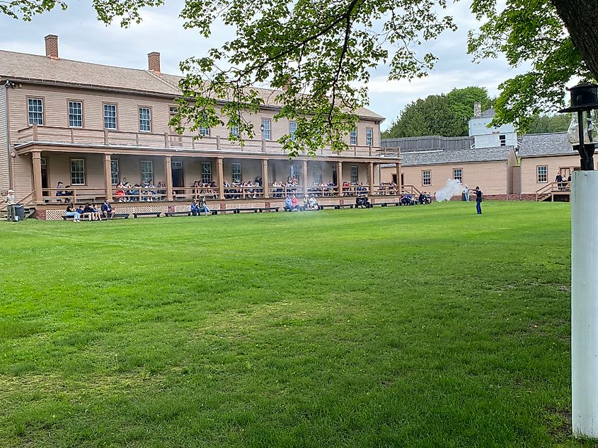A scout in an American Revolutionary War outfit demonstrates a rifle firing in front of a small crowd seated on the adjacent army barracks