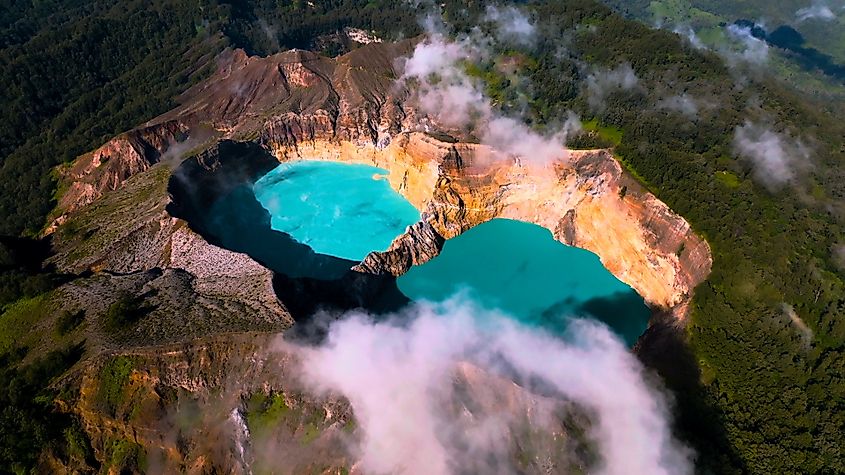 Aerial view of Kelimutu volcano with its three colorful crater lakes surrounded by lush greenery and clouds.