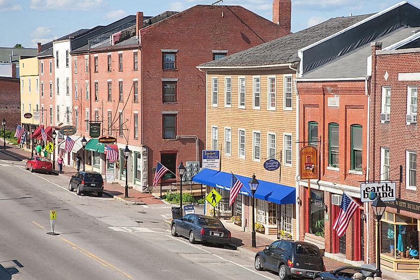 A quaint American small-town street with brick buildings adorned with U.S. flags, colorful storefronts, and parked cars under a bright blue sky.