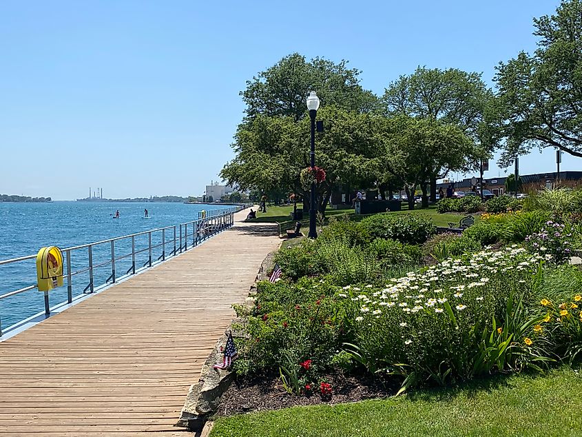 A riverside boardwalk with beds of flowers and passing paddleboarders.