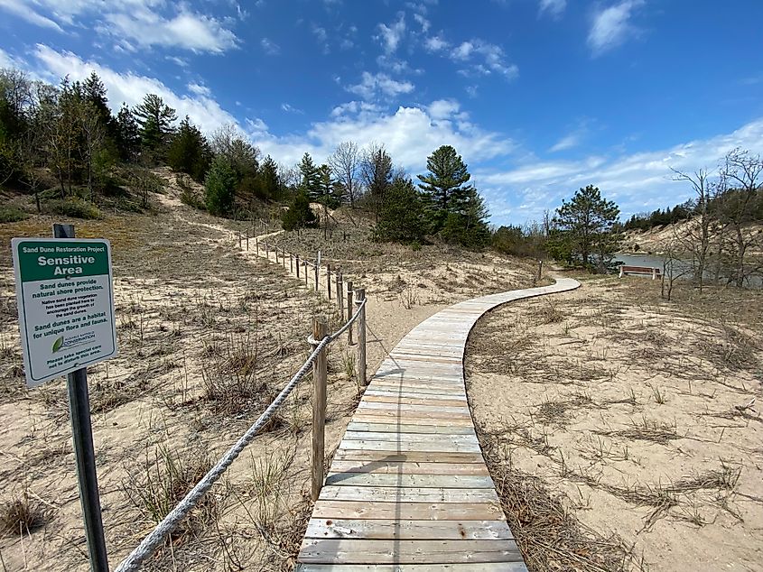 A boardwalk leads through a set of riverside sand dunes marked by a small green sign as "Sensitive Area."