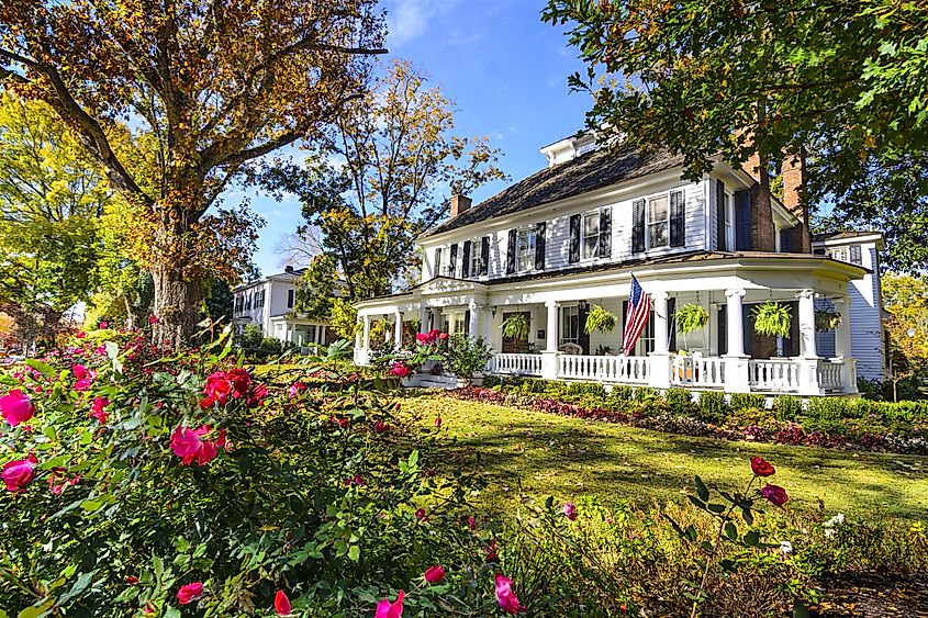 Charming white colonial house with wraparound porch, lush garden, red roses, and American flag. Autumn trees frame the scene under clear blue sky.