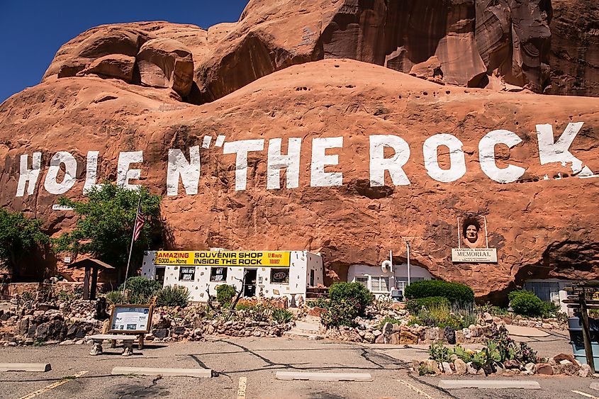 Massive red rock with "Hole N' The Rock" in white letters. Below, a souvenir shop and memorial. Bright day, blue sky, and desert plants.
