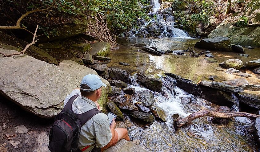 Man with backpack sitting on the rock reading tablet by the waterfall.