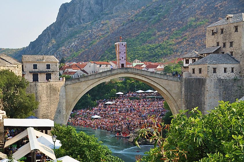 Thousands of riverside spectators watching the Red Bull diving series. A large platform has been set up atop the Old Bridge, and a brave diver is ready to make the leap into the river.  