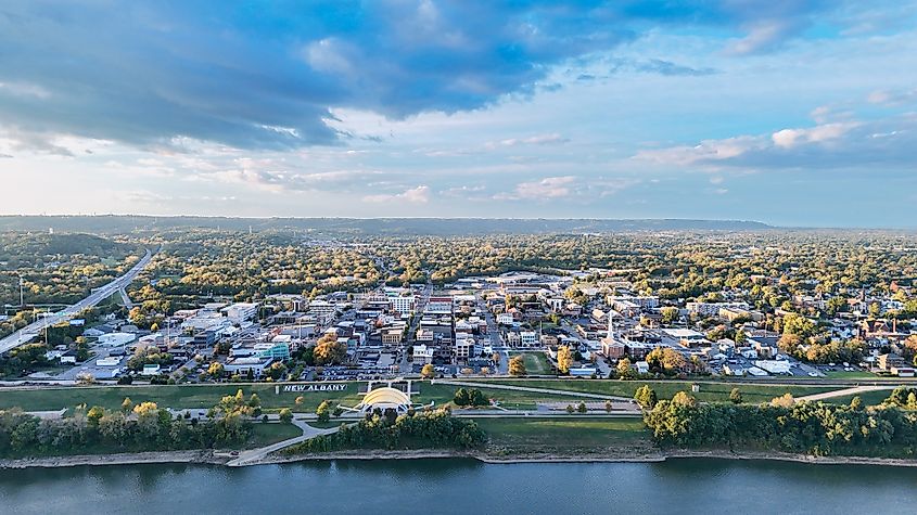 A wide aerial view of downtown New Albany, Indiana, shows the city stretching along the Ohio River. A riverfront park and amphitheater sit beside the water, while streets lined with buildings, homes, and trees extend into the distance under a partly cloudy sky.