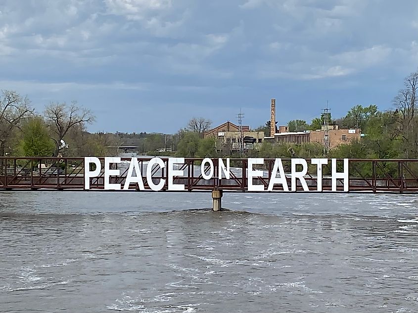 A rust colored pedestrian bridge with white block letters reading 'Peace on Earth' crosses a wide river.