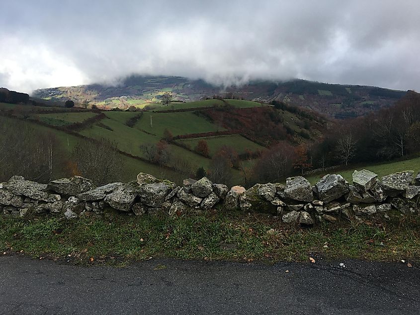 A small stone wall portions off a concrete path from the surrounding hilly, verdant countryside. It is a gloomy day. 