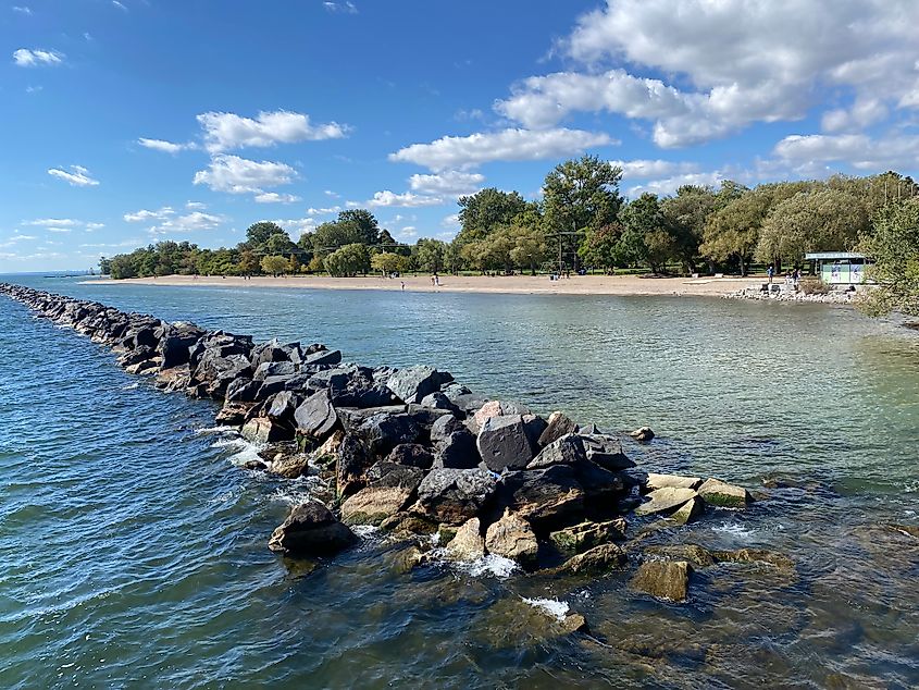A rocky breakwall in the middle of blue waters before a long sandy beach.