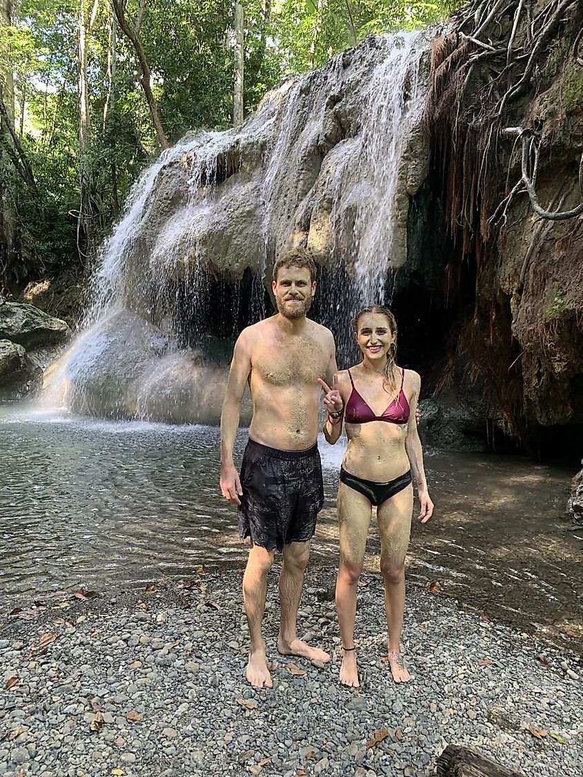 A mud-covered couple in bathing suits stand in front of a waterfall