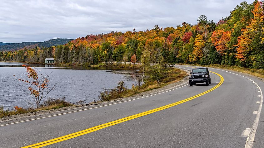 SUV driving along Beaver Pond on winding Route 17, part of Rangeley Lake Scenic Byway in Rangeley, Maine, USA, on a cloudy but colorful autumn morning.