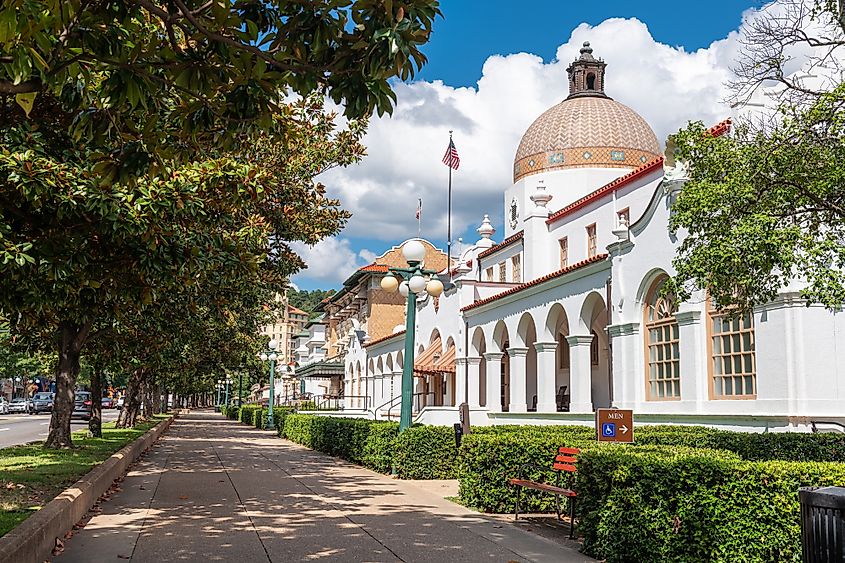 A historic white building with a domed roof and arches, lined with green hedges and trees. A clear blue sky and clouds create a peaceful atmosphere.