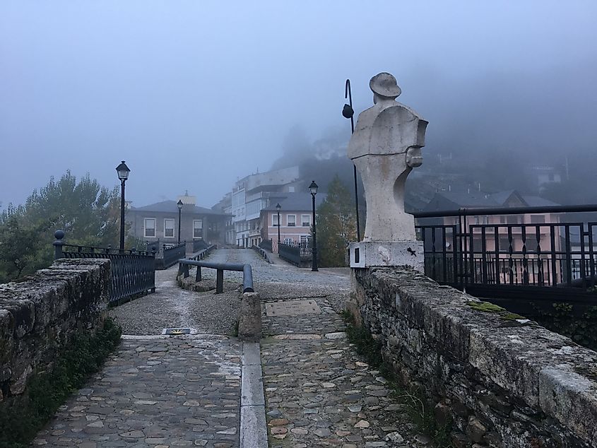 On a foggy afternoon, a marble pilgrim statue guards a medieval bridge
