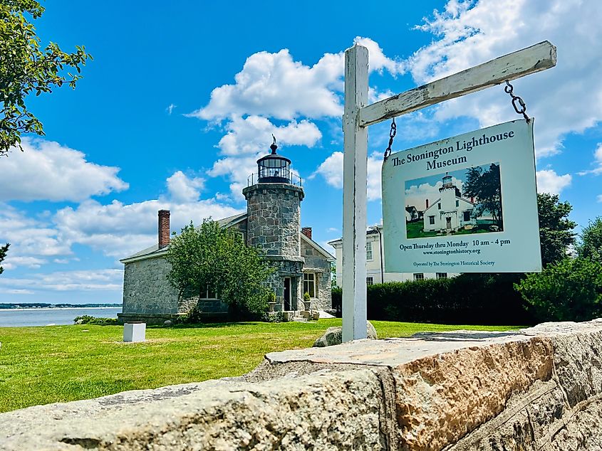 Stone lighthouse in Stonington Borough, Connecticut, with a sign in the foreground and water visible in the background under a blue sky.