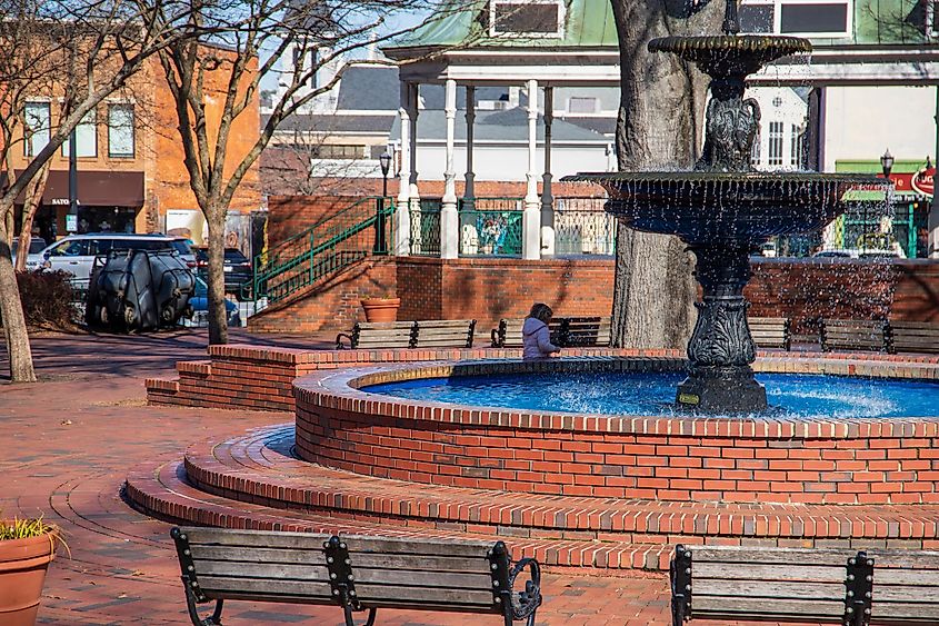 A circular red brick water fountain with benches, bare winter trees and a little girl running and parked cars at the Marietta Square in Marietta Georgia USA.