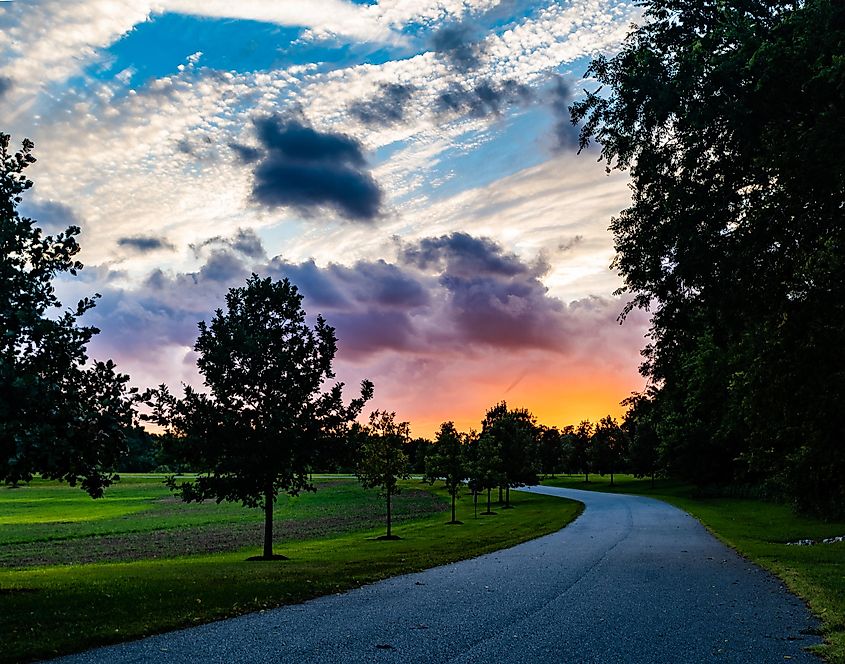 A path leads through trees forest and meadow into a brilliant orange and yellow sunset beneath menacing storm clouds.