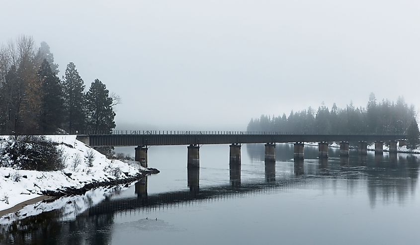 A railroad bridge crosses over the calm Clark Fork River on a foggy winter day in December in north Idaho.