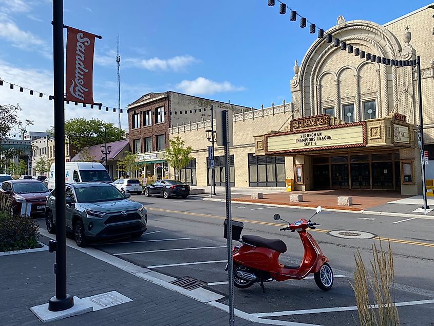 A small red scooter parked below a red "Sandusky" sign, outside of a stately theatre.