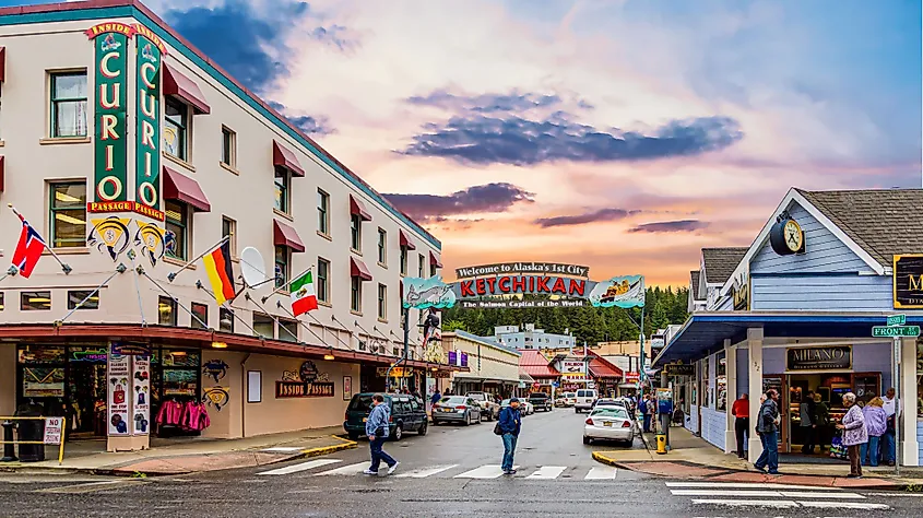 Busy street in Ketchikan, Alaska, at sunset. Colorful sky, people walking, shops with various flags, and a welcoming sign create a lively, inviting atmosphere.
