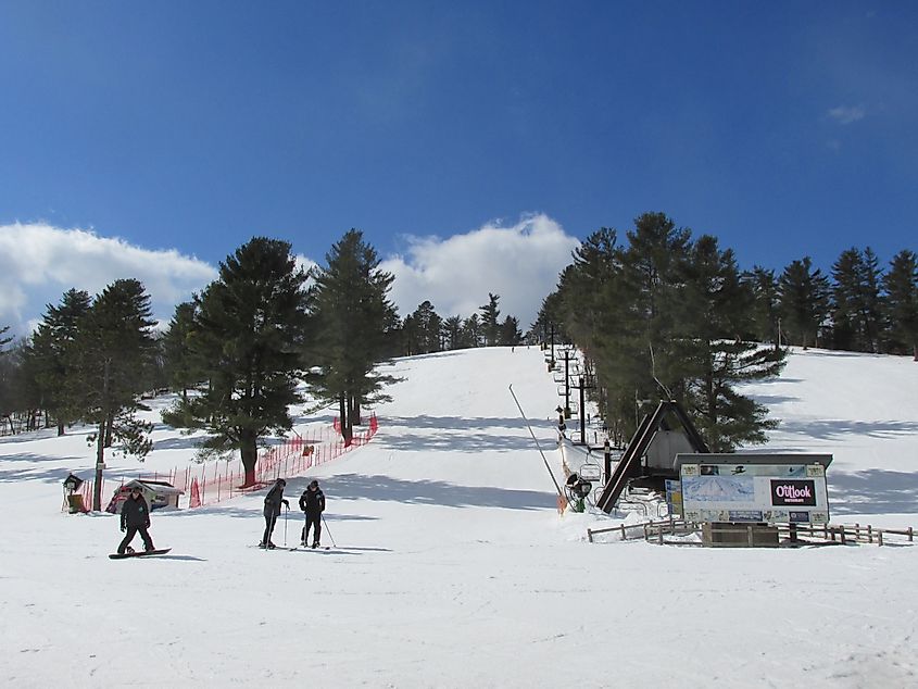Ski resort on a sunny day, featuring a snow-covered slope with skiers near the bottom. Pine trees lining the piste, with clear blue sky above.