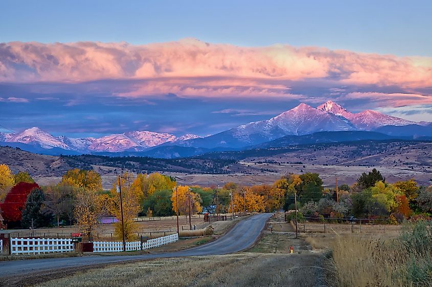 Long's Peak lights up at sunrise as a rural country road leads into the fall trees