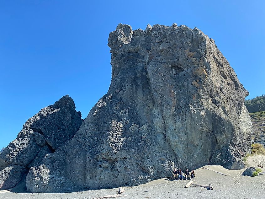A group of four sit in the sand, leaning up against a massive rock formation.