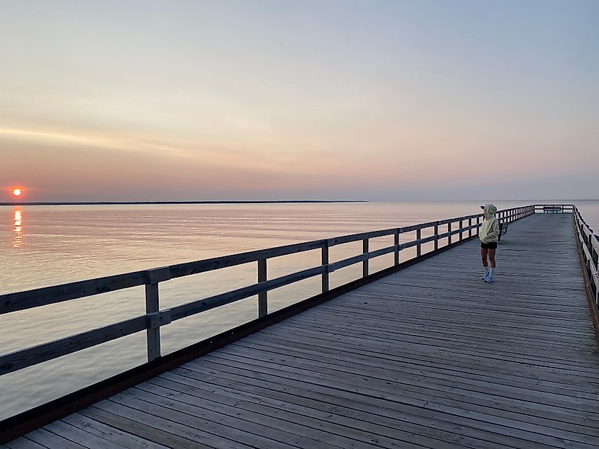A woman in a yellow windbreaker looks toward sunrise while walking a lakeside pier.