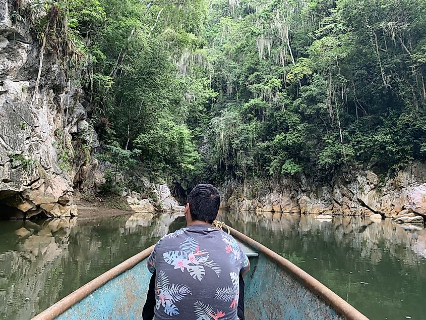 A man sits in the front of a paddle boat that is heading into a lush canyon