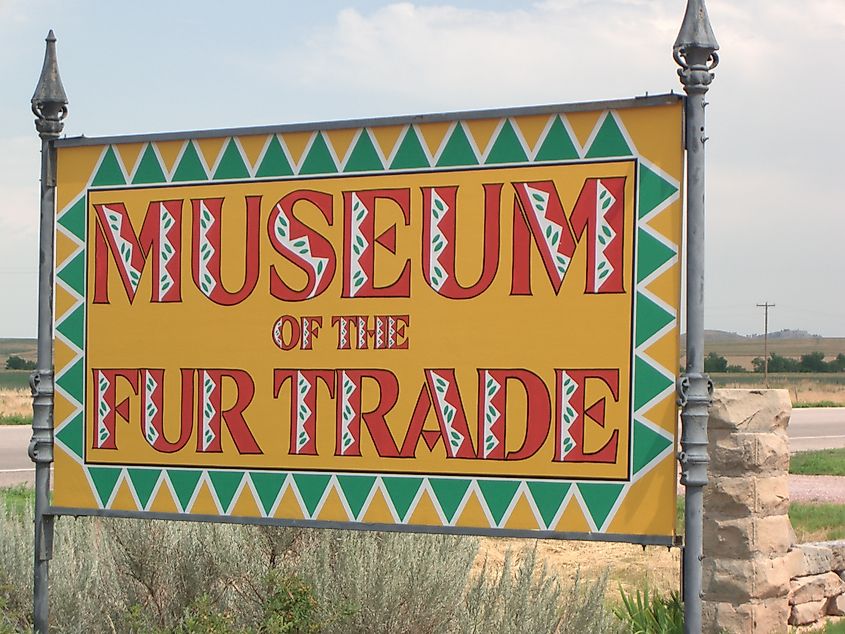 Sign for "Museum of the Fur Trade" with red text on a yellow background, bordered by green and yellow geometric patterns, under a partly cloudy sky.