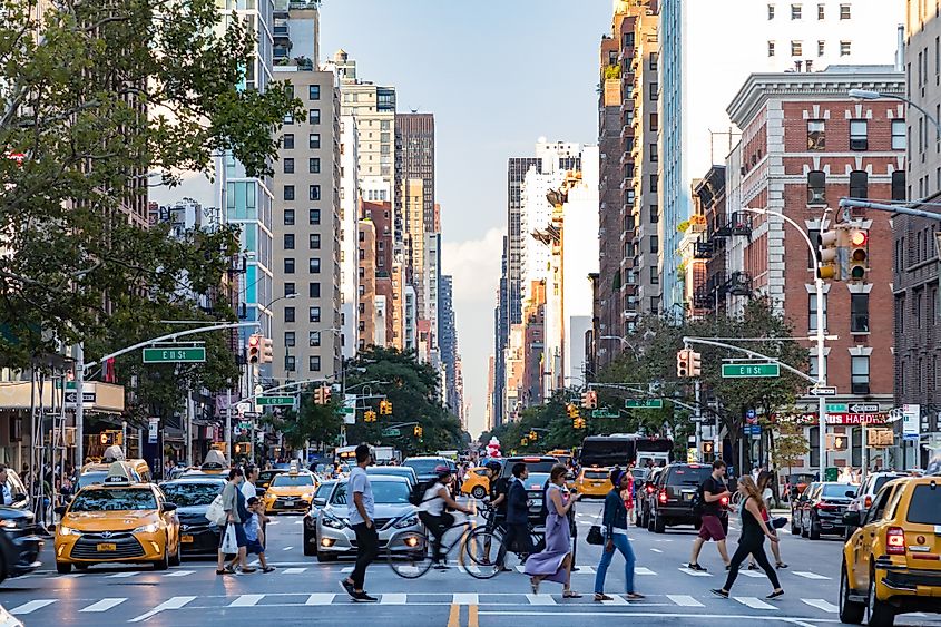 Busy crowds of people walk across 3rd Avenue in front of rush hour traffic in the East Village neighborhood of Manhattan in New York City.