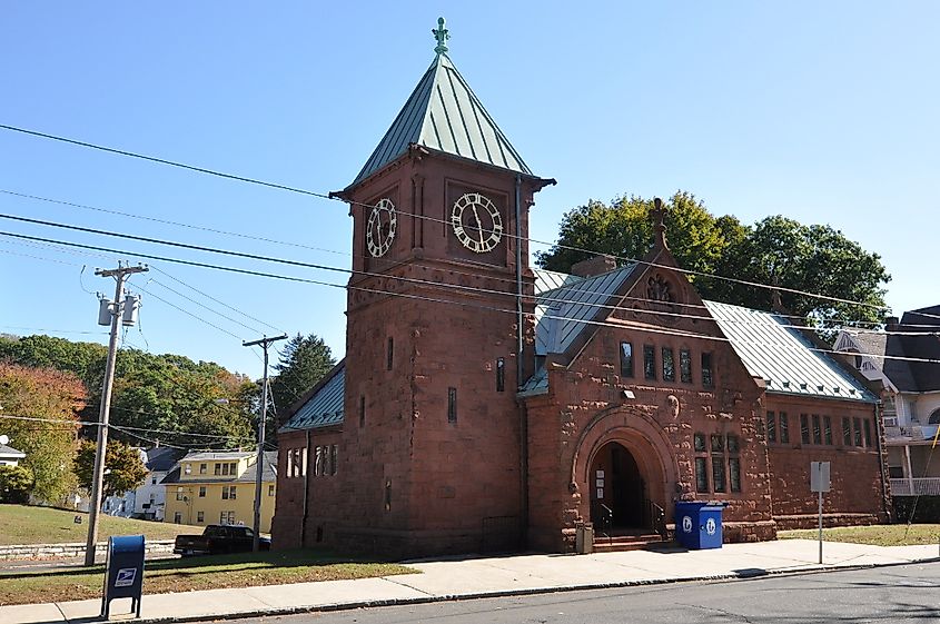 The Ansonia Library has a clock tower and green roof accents, situated beside a street with overhead power lines, neighboring houses, and a blue mailbox in the foreground.