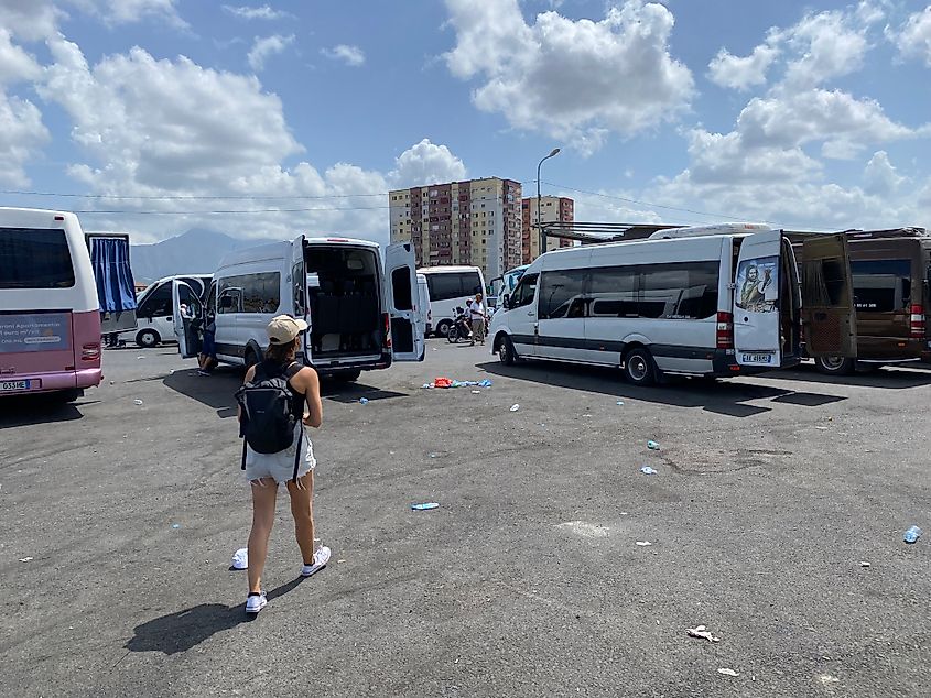 A female backpacker walks through a littered parking lot with various shuttle buses.