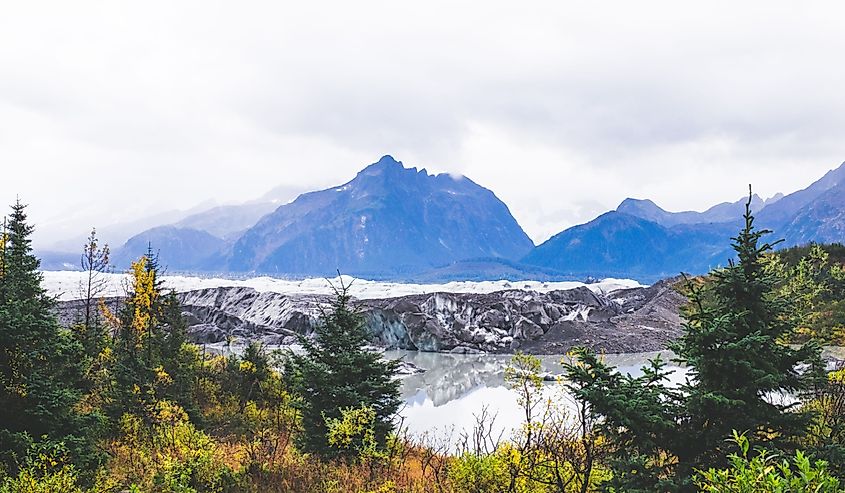 Scenic view of a glacier with reflective water surrounded by lush greenery. Majestic mountains rise in the background under a cloudy sky, evoking tranquility.