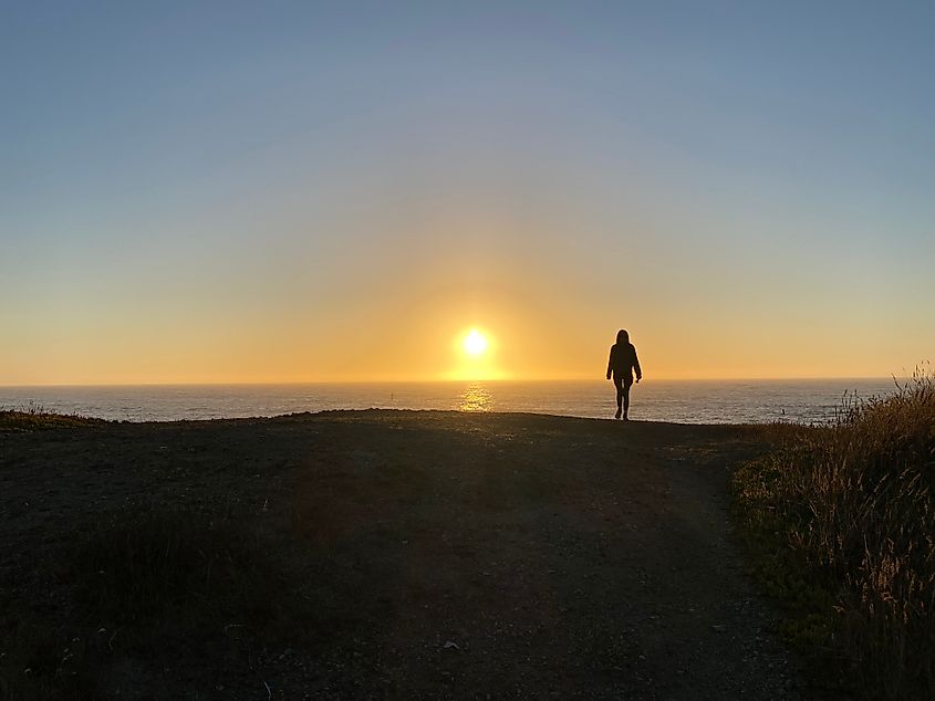 The silhouette of a woman standing on a seaside hilltop as the sun sets.