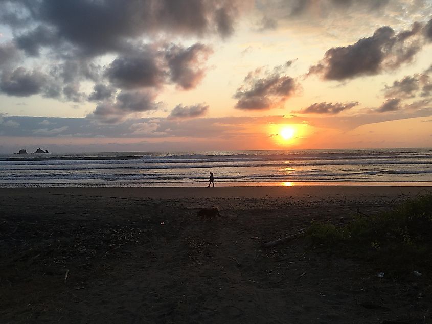 A man walks alone on an empty beach while the sun sets in the background