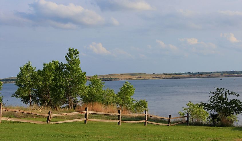 A shot of Kanopolis Lake on the north side of a Wooden Fence, green grass,water,trees, blue sky and clouds, that is bright and colorful