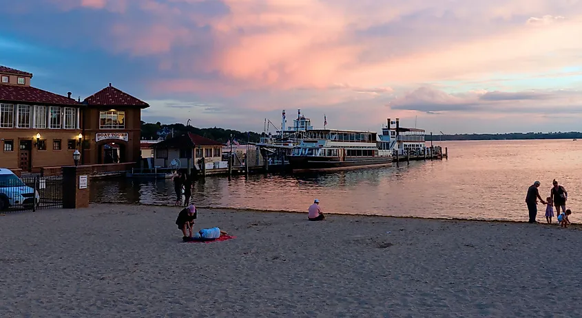 People gathering at the beach near Lake Geneva at sunset, with boat docks in view.